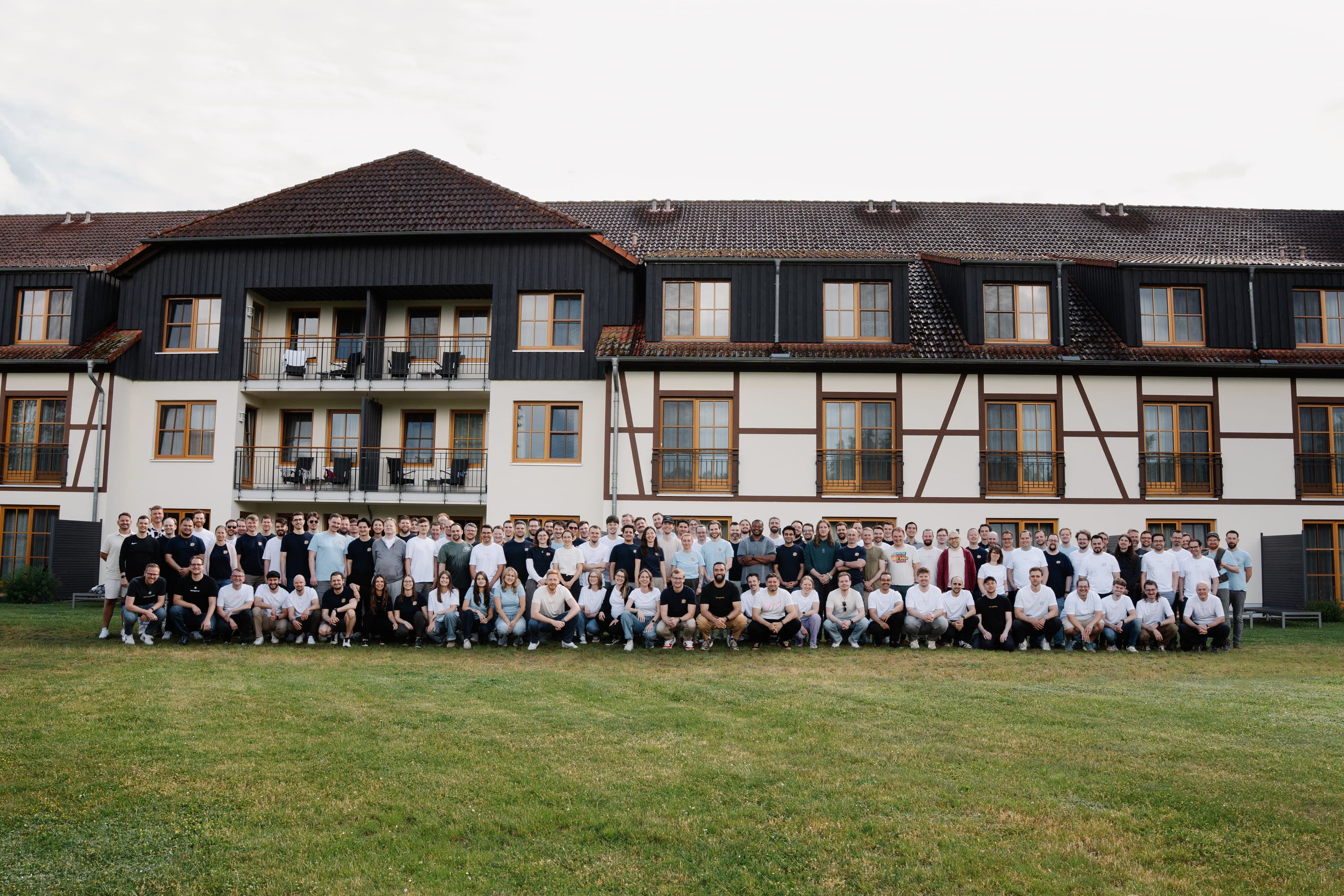 A large group of around 100 iits employees posing for a team photo on a grassy lawn in front of a traditional half-timbered building.